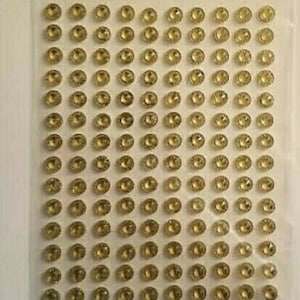 May include: A sheet of gold self-adhesive rhinestones. The rhinestones are arranged in a grid pattern on a clear plastic sheet.