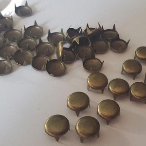 May include: A large quantity of gold-toned metal studs with a rounded top and three prongs on the bottom. The studs are scattered on a white surface.