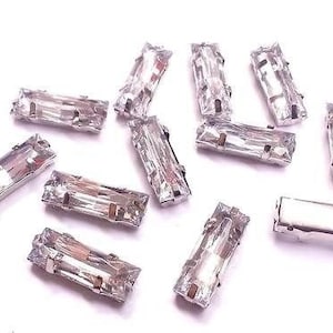 May include: A collection of ten clear crystal rhinestones in a rectangular shape. The rhinestones have a silver backing and are arranged on a white surface.