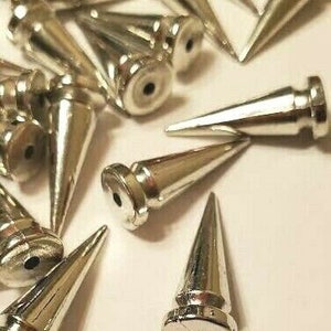 Pode incluir: Um close-up de um grupo de tachas piramidais de metal prateado. As tachas são todas do mesmo tamanho e forma, e têm uma ponta pontiaguda. Elas estão dispostas em um padrão aleatório em uma superfície branca.