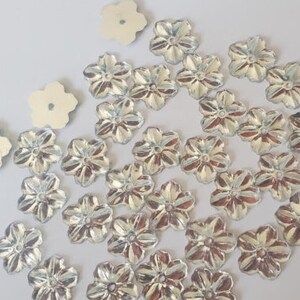 May include: A collection of silver-colored flower-shaped embellishments. The embellishments have a raised design and a hole in the center.
