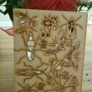 May include: A wooden plaque with a laser-cut design of various insects, including butterflies, beetles, and ladybugs. The insects are arranged in a whimsical pattern on a light brown background.