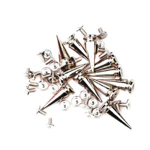 May include: A pile of silver metal cone studs with pointed ends and screw fixings. The studs are scattered on a white background, with several small screws and washers also visible.