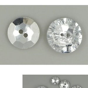 Può includere: Due bottoni rotondi argentati con superfici sfaccettate e due fori ciascuno. I bottoni hanno una qualità lucida e riflettente, che suggerisce un materiale metallico o cristallino. Sono posti su uno sfondo grigio chiaro.