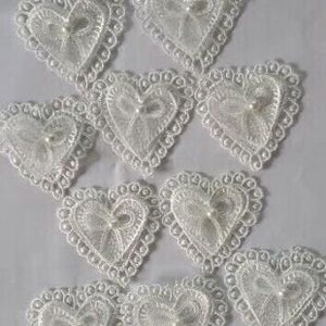 Puede incluir: Diez adornos de encaje blanco en forma de corazón con una perla en el centro de cada corazón.