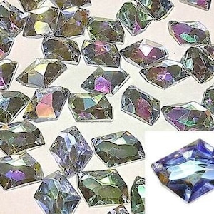 Pode incluir: Uma grande quantidade de gemas de vidro retangulares facetadas e iridescentes. As gemas estão espalhadas sobre uma superfície branca.