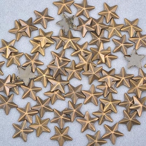 May include: A collection of bronze-colored star-shaped metal embellishments. The stars have a textured surface and are scattered across a light gray background. Several stars have a silver-colored finish.