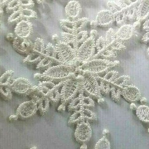 May include: White lace fabric with a snowflake design. The lace is delicate and intricate, with a floral pattern.