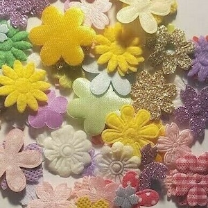 May include: Assortment of colorful felt flower embellishments in various shapes and sizes. The flowers are made of felt and have a variety of textures, including smooth, fuzzy, and textured. The flowers are in shades of pink, purple, yellow, green, and white.