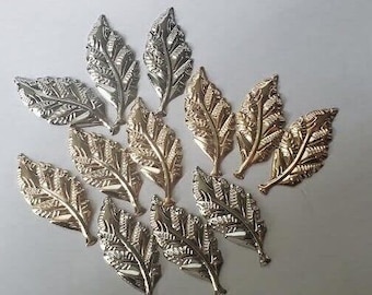 May include: An assortment of decorative metal leaf embellishments in silver and gold tones. The leaves have detailed veining and are arranged on a white surface. These are suitable for craft projects.