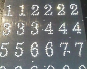 May include: A sheet of self-adhesive rhinestone numbers, featuring the numbers 0 through 9. Each number is made of small, clear rhinestones arranged in a numeral shape.