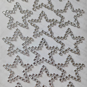 Op de afbeelding: Een verzameling stervormige strass-versieringen. Elke ster is omrand met kleine, heldere strass-steentjes, wat een sprankelend effect creëert. De sterren zijn gerangschikt in een herhalend patroon op een wit oppervlak, geschikt voor knutselen en decoraties.