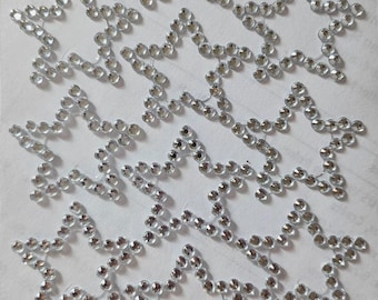 May include: A collection of star-shaped rhinestone embellishments. Each star is outlined with small, clear rhinestones, creating a sparkling effect. The stars are arranged in a repeating pattern on a white surface, suitable for crafts and decorations.