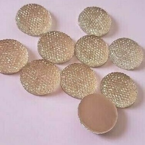 May include: A collection of round, decorative embellishments. The majority of the embellishments are a light gold color with a textured, sparkling surface. One embellishment is a solid, light brown color. The objects are arranged on a light pink surface.
