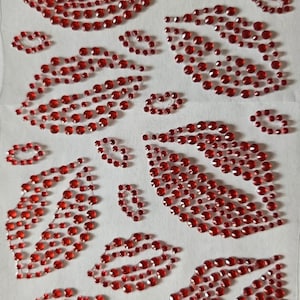 Op de afbeelding: Vel rode strass steentjes lip- en kusontwerpen. De ontwerpen zijn gerangschikt in verschillende maten en oriëntaties, waarbij de strass steentjes een sprankelend effect creëren. De vellen liggen op een witte achtergrond.