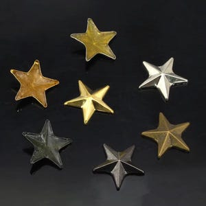 May include: A collection of seven five-pointed star-shaped studs in various metallic finishes, including polished gold, silver, antique bronze, and dark gunmetal, arranged in a scattered pattern against a dark background.