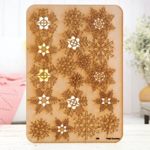 May include: A sheet of wood with a laser-cut design of snowflakes. The snowflakes are of various sizes and shapes, and some have intricate details. The wood is a light brown color.