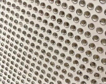 May include: Close-up of a white surface densely covered with numerous, evenly spaced, clear, faceted, round rhinestones. The rhinestones reflect light, creating a sparkling effect. The arrangement suggests a decorative or craft element.