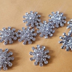 May include: A collection of silver-colored snowflake-shaped embellishments, each encrusted with numerous clear, sparkling rhinestones. The embellishments are arranged on a light brown surface, showcasing their intricate design.