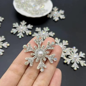 May include: Close-up of several white snowflake embellishments with iridescent rhinestones and a pearl center. Some snowflakes are scattered on a dark surface, while others are in a small white dish. These decorative accents are ideal for crafts and holiday projects.