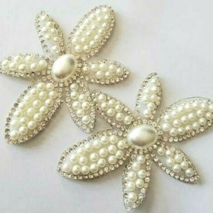 May include: Two decorative flower appliques. Each flower has six petals, each petal is filled with small, white, faux pearls. The petals are outlined with clear rhinestones. The center of each flower is a large, oval, pearl-like bead.
