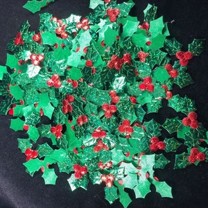 May include: A large pile of green glitter holly leaves with red glitter berries. The leaves and berries are scattered on a black background.