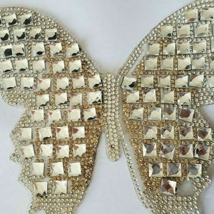 May include: A silver and gold butterfly-shaped rhinestone applique. The applique is made of small, square rhinestones that are arranged in a pattern. The applique is perfect for adding a touch of sparkle to any project.