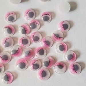 May include: A collection of plastic googly eyes with pink eyelids and black pupils. The eyes are various sizes and are scattered on a white surface.