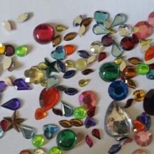 May include: Assortment of colorful rhinestones in various shapes and sizes, including stars, hearts, teardrops, and ovals. The rhinestones are scattered on a white surface.