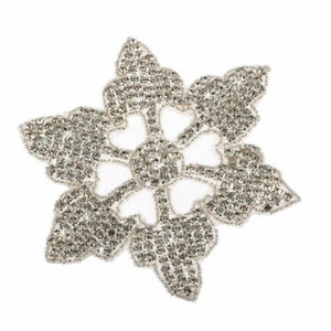 May include: A silver snowflake-shaped embellishment with a sparkly, crystal-like finish. The snowflake is made of many small, individual crystals that are sewn together to create a delicate and intricate design.