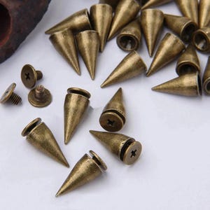 May include: A collection of antique brass-coloured metal cone-shaped spikes. Each spike features a pointed top and a flat circular base with a threaded screw attachment. The finish has a weathered, metallic texture with visible cross-head screws.