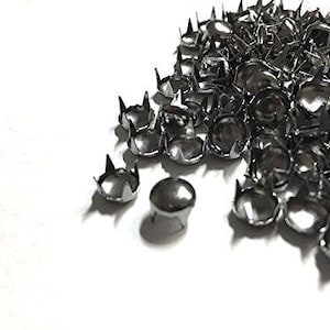 May include: A pile of small, silver-colored metal studs with a rounded top and four pointed prongs. The studs are scattered on a white surface, with some casting shadows. These decorative studs are likely used for embellishing clothing or accessories.