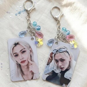 May include: Two rectangular keychains featuring portraits of a person with blonde hair. Each keychain has a silver metal lobster clasp and a chain adorned with small, colorful translucent beads in blue, pink, and yellow star shapes.