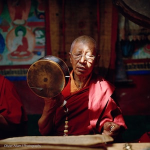 Puede incluir: Un monje budista con una túnica roja sostiene un tambor de mano marrón y recita una oración. Lleva gafas y tiene una expresión seria en su rostro.