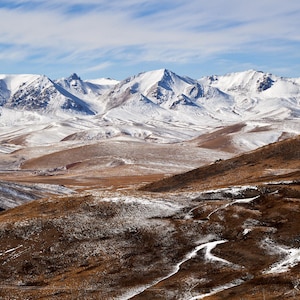Pode incluir: Uma paisagem cénica com montanhas cobertas de neve sob um céu azul com nuvens dispersas. O primeiro plano mostra colinas marrons e gramadas com manchas de neve. A imagem captura um ambiente natural sereno.