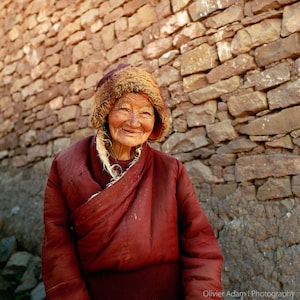 Puede incluir: Una mujer mayor con una cálida sonrisa, vestida con una túnica roja tradicional y un gorro de piel marrón, se encuentra frente a una pared de piedra.