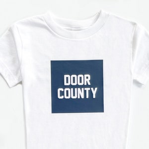 May include: White t-shirt with a blue square on the front that says "DOOR COUNTY" in white letters.