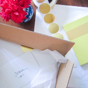May include: A brown cardboard box with a white tissue paper lining. The box is open and a white card with the word "Shana" written on it is inside. The box is sitting on a white surface with a yellow sticky note and a sheet of paper with gold circles.
