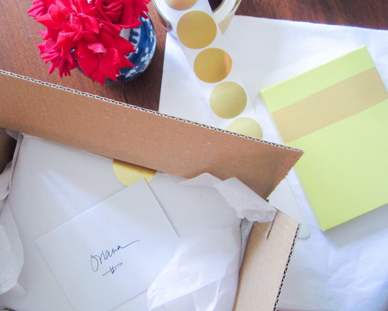 May include: A brown cardboard box with a white tissue paper lining. The box is open and a white card with the word "Shana" written on it is inside. The box is sitting on a white surface with a yellow sticky note and a sheet of paper with gold circles.