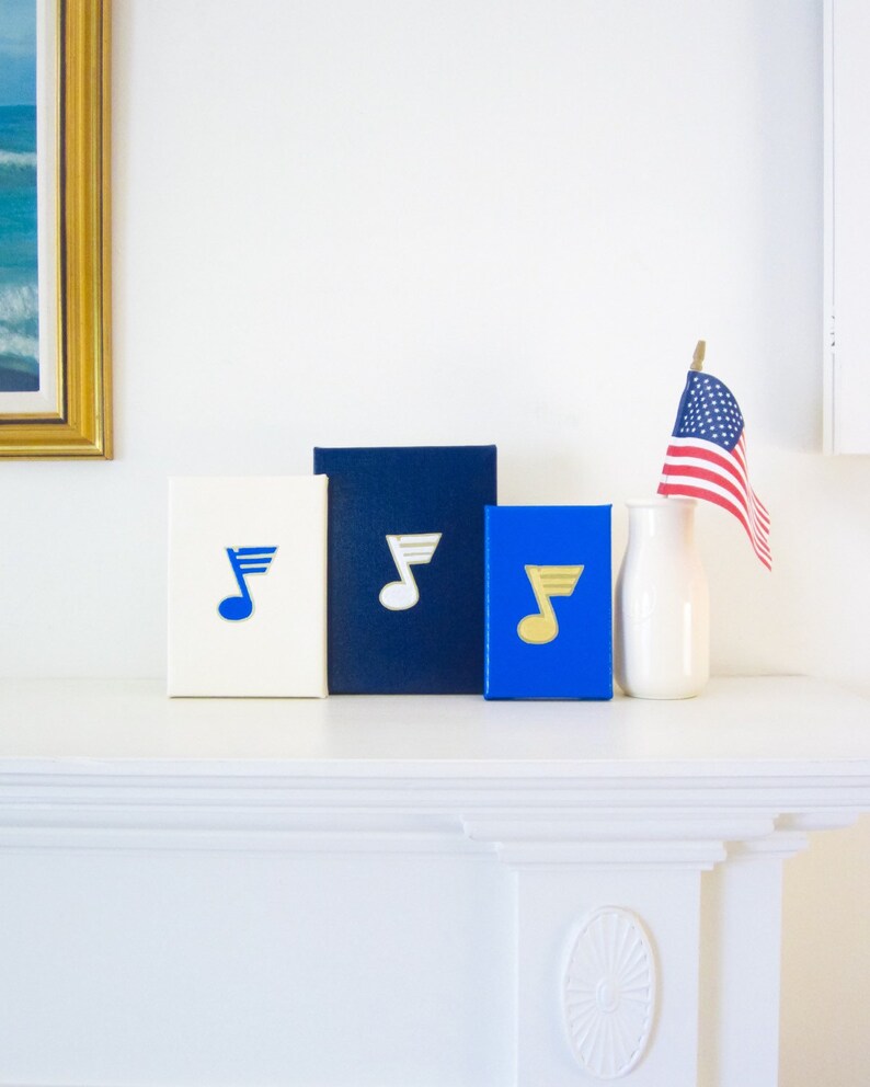 May include: Three decorative boxes with a musical note design. The boxes are white, navy blue, and royal blue. The musical notes are gold, white, and blue. The boxes are on a white mantel.