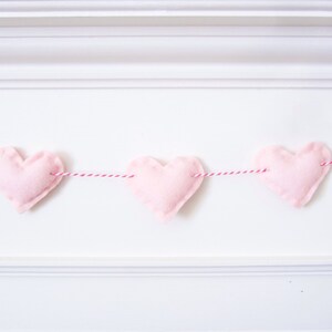 Puede incluir: Una guirnalda de corazones de fieltro rosa con tres corazones enhebrados en un cordel rojo y blanco.