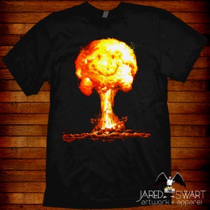 May include: A black t-shirt featuring a graphic of a smiling mushroom cloud explosion. The graphic is in shades of orange, yellow, and red.