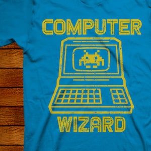 Peut inclure: Un t-shirt bleu avec un graphique jaune d'un ordinateur rétro avec le texte "COMPUTER WIZARD" au-dessus.