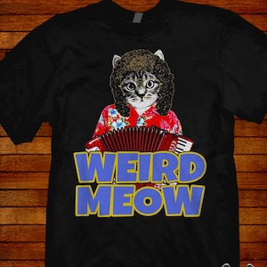 May include: Black t-shirt featuring a cat with glasses, a curly wig, and a Hawaiian shirt, playing an accordion. The text "WEIRD MEOW" is in blue with a yellow outline. The artist's signature is at the bottom.