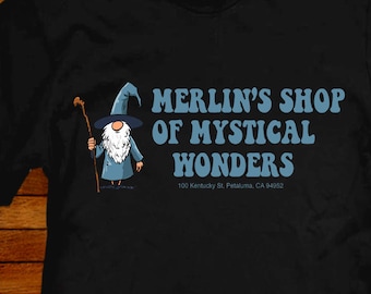 Può includere: Maglietta nera con un mago dei cartoni animati e il testo "Merlin's Shop of Mystical Wonders" e l'indirizzo "100 Kentucky St. Petaluma, CA 94952".