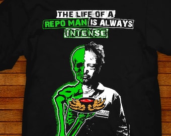 May include: Black t-shirt with a green and white illustration of a skeleton holding a bowl with red liquid. The text "THE LIFE OF A REPO MAN IS ALWAYS INTENSE" is printed in white on the shirt. The text "MILLER" is printed in white on the illustration. The text "JARED SWART artwork + apparel" is printed in white on the bottom of the shirt.