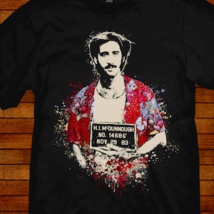 May include: Black t-shirt with a colorful graphic of a man holding a mugshot. The mugshot reads "H.I. M-DUNNOUGH NO. 14686 NOV 29 83".