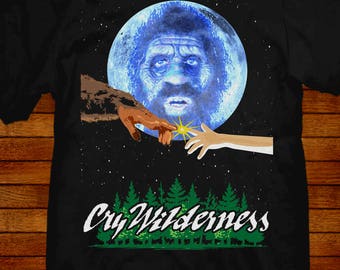 Può includere: Maglietta nera con una luna blu e una scena di foresta. La luna ha un viso e due mani si tendono per toccarla. Il testo "Cry Wilderness" è stampato sulla maglietta.
