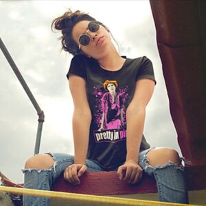 May include: A woman wearing a black t-shirt with a pink and white graphic of a woman with the text "pretty in pink" on the front. She is wearing blue jeans with a ripped knee and round sunglasses.
