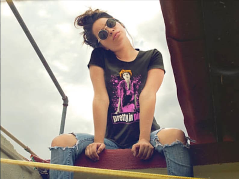 May include: A woman wearing a black t-shirt with a pink and white graphic of a woman with the text "pretty in pink" on the front. She is wearing blue jeans with a ripped knee and round sunglasses.